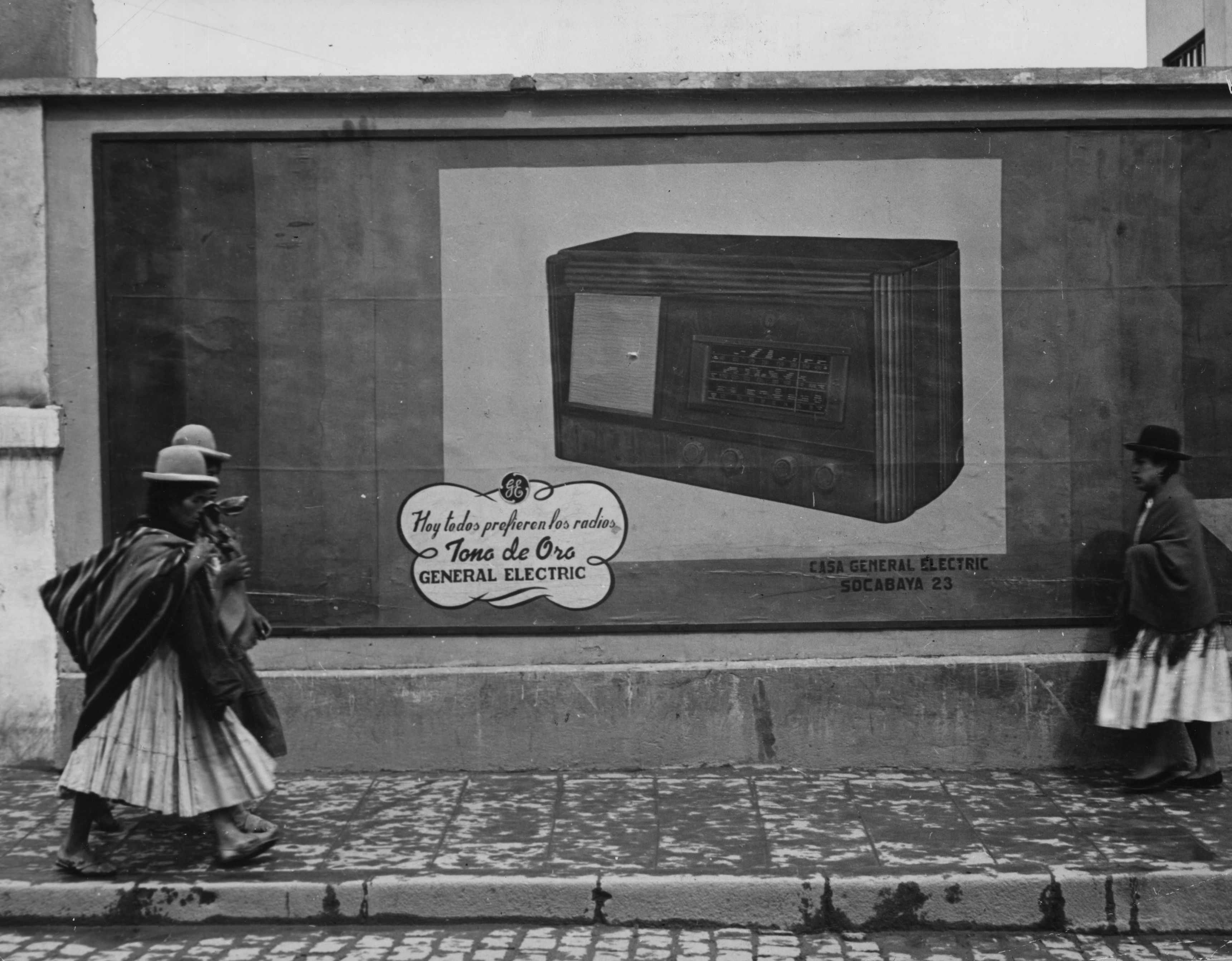 Black & White photo of Bolivian women in traditional clothes passing in front of a shop. 